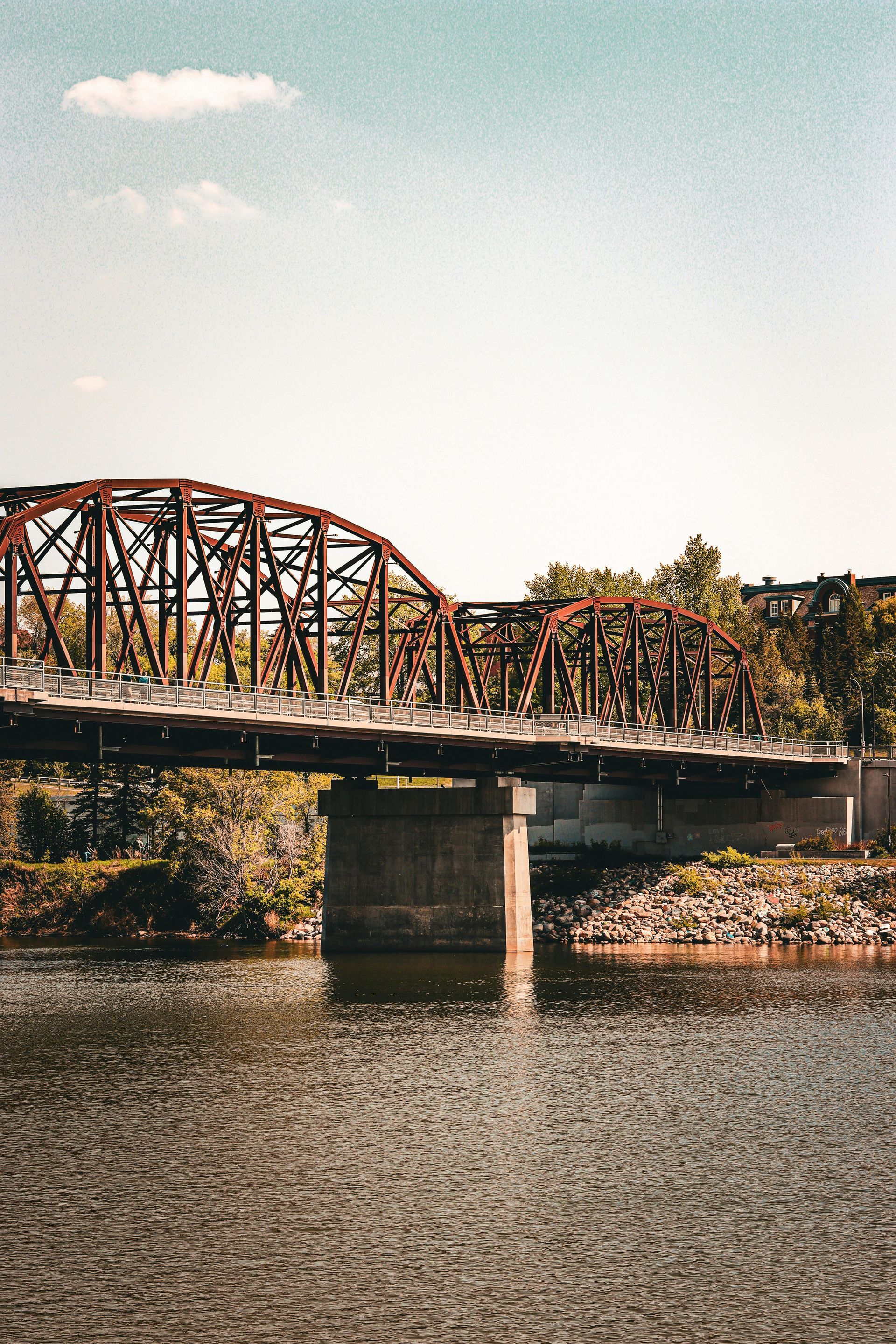 Rusty truss bridge over a river on a sunny day, with trees along the bank