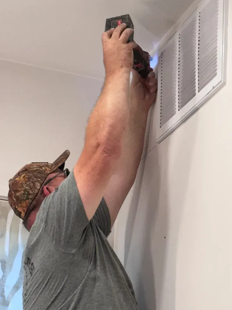 A man is using a drill to fix an air vent on a wall.