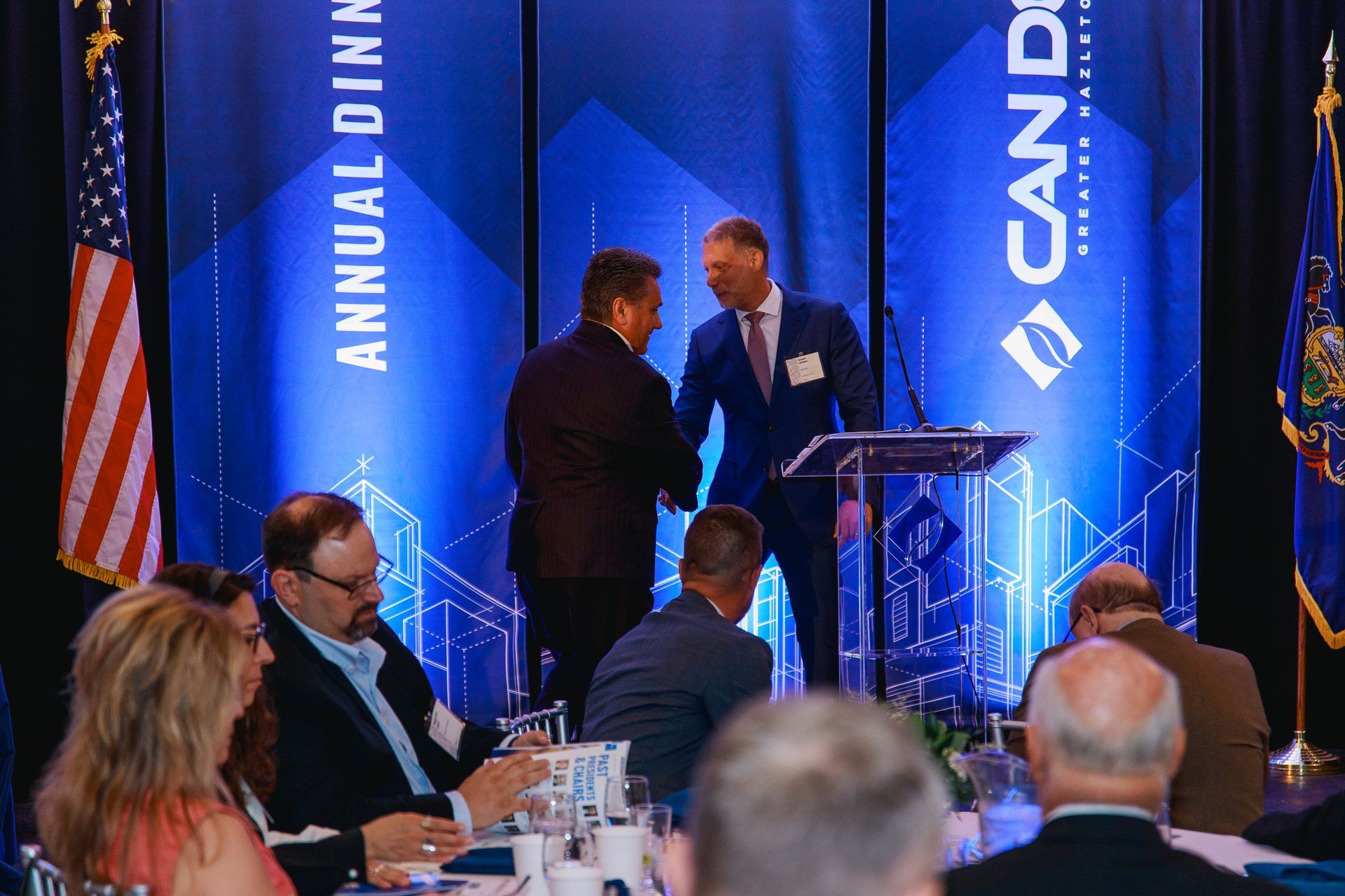 Two men shaking hands on a blue-lit conference stage with a podium and audience in the foreground.