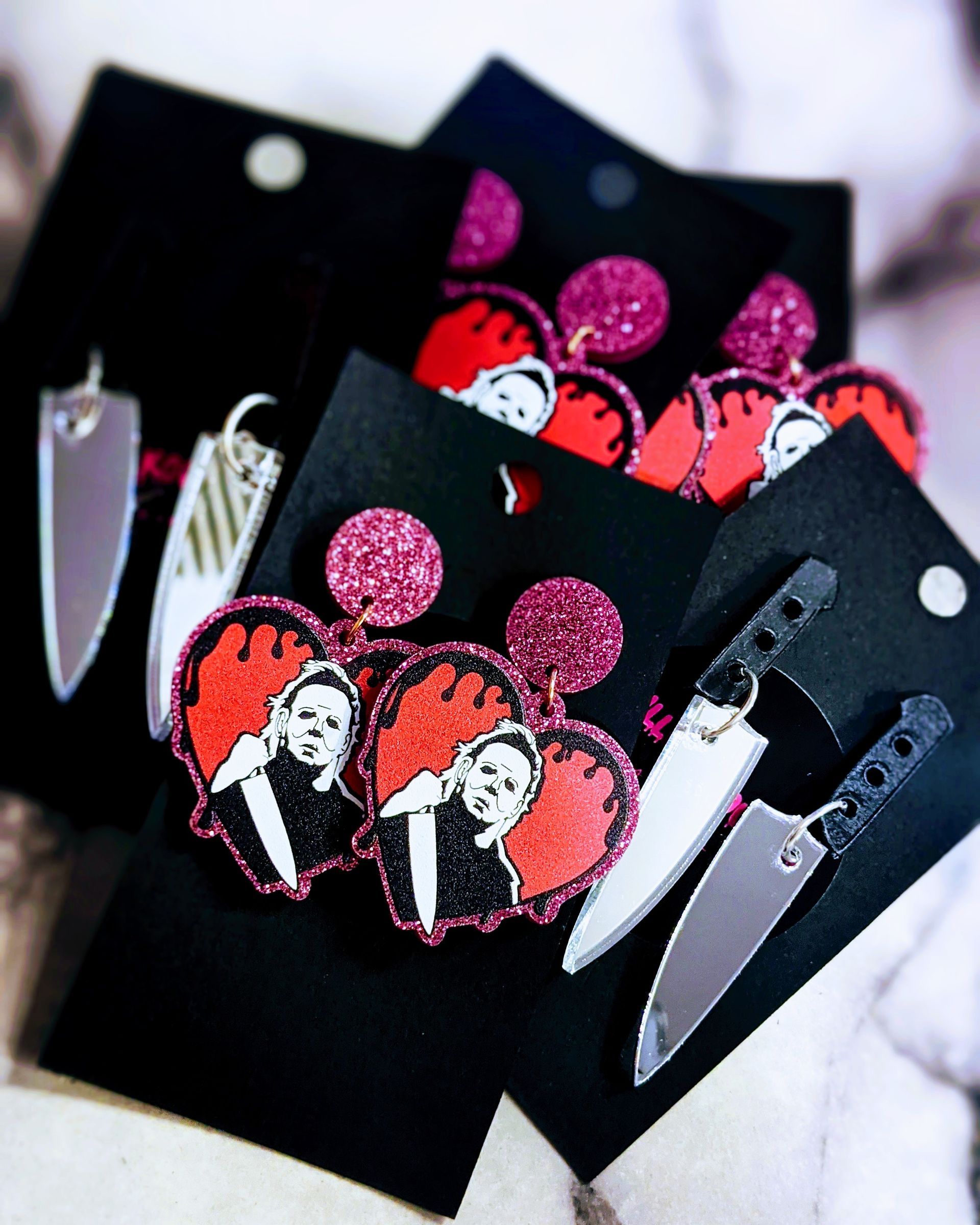 Multiple pairs of horror-themed earrings, featuring Michael Myers heart-shaped charms and silver knife designs on cards.