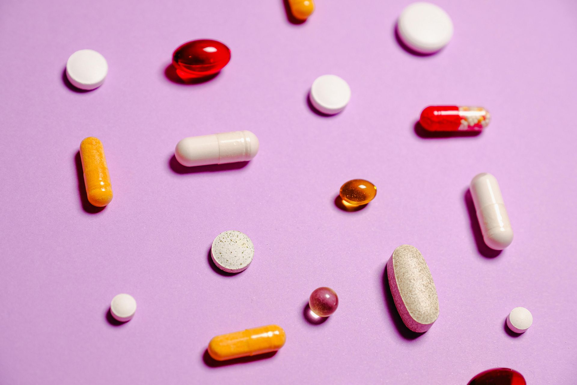 Various pills, capsules, and softgels of different shapes and colors scattered on a plain light purple background.