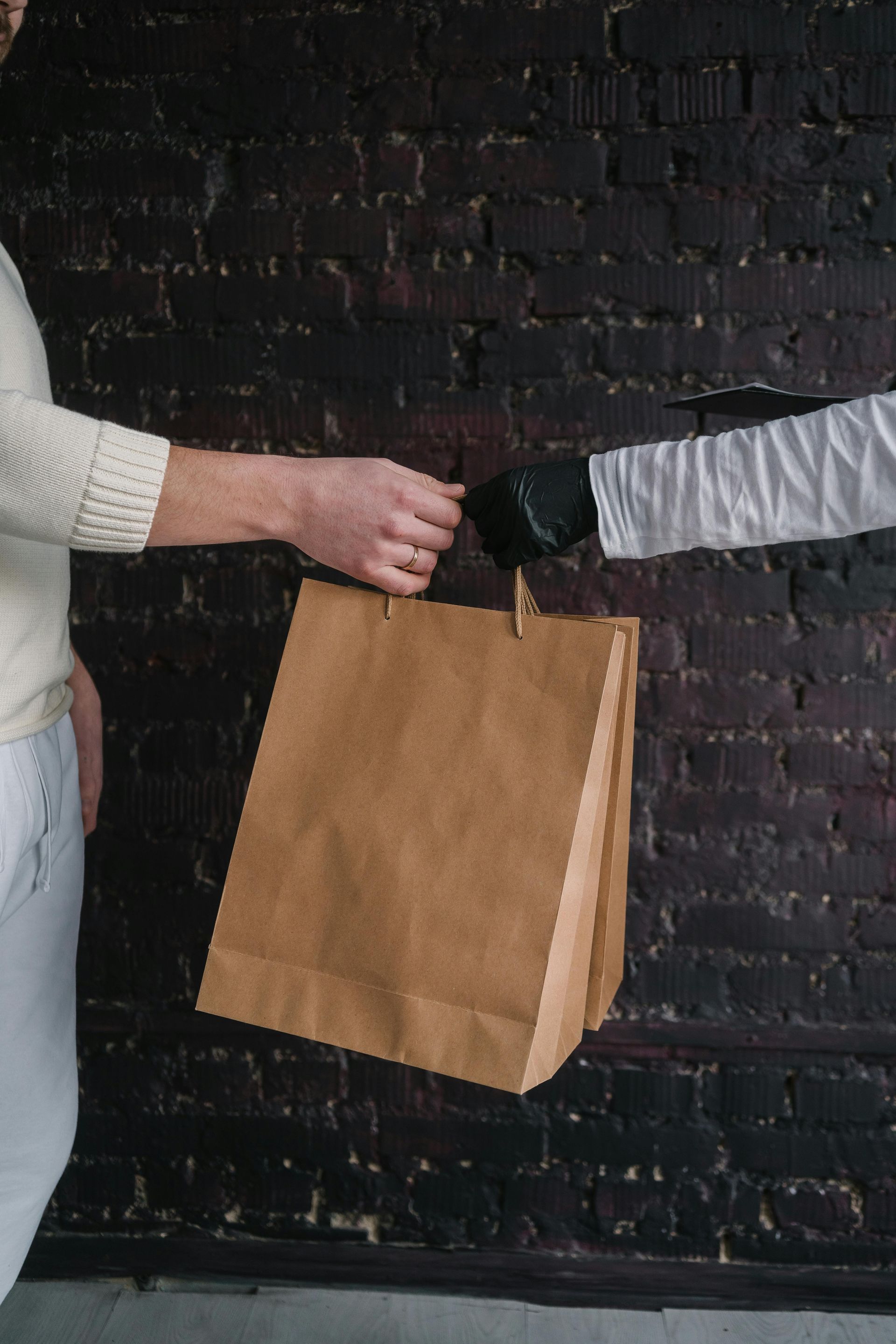 A person in a white sweater takes a brown paper bag from a hand wearing a black glove against a dark brick background.