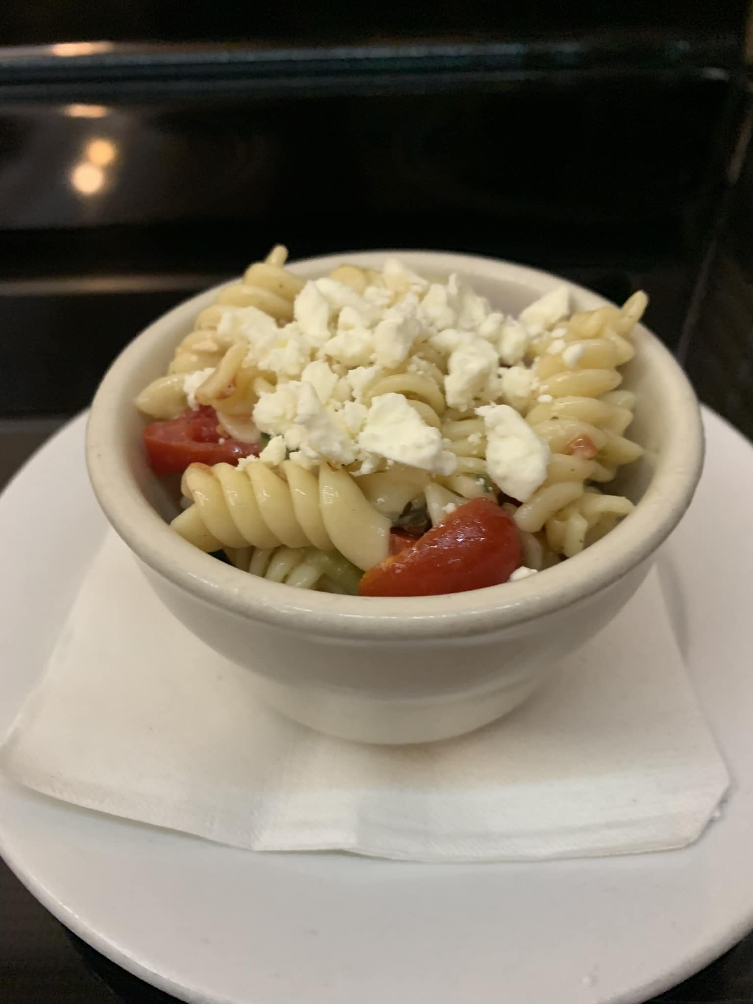 Pasta salad in a white bowl, topped with feta cheese and tomatoes, on a white plate.