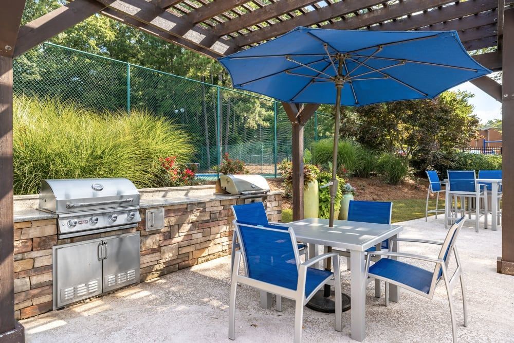 Outdoor table and chairs under a pergola with a blue umbrella right next to the grilling station at Marquis at Perimeter Center in Atlanta, GA.