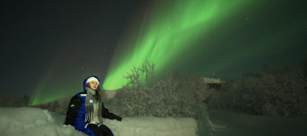 girl standing under aurora sky and northern lights colored in red and green sitting on snow dressed in winter clothes
