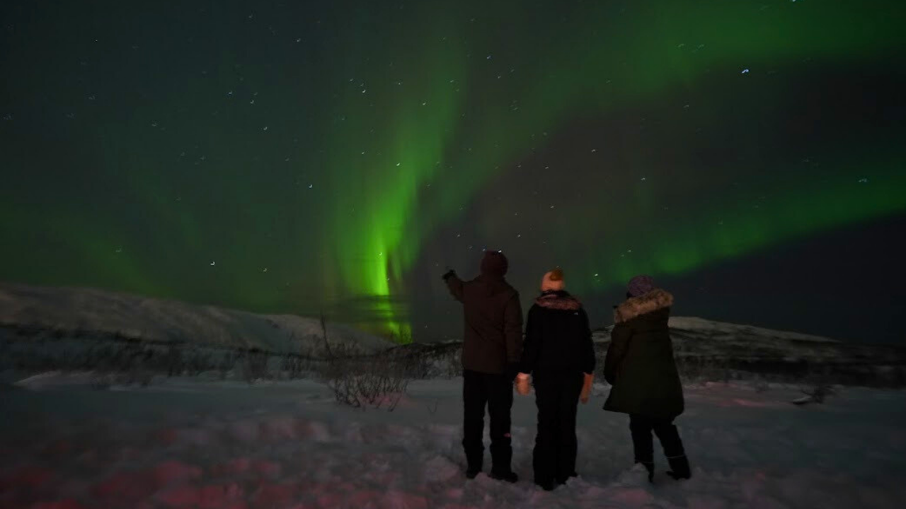 two women one man standing infront of aurora northern light view dressed in winter clothing and the ground is covered in snow