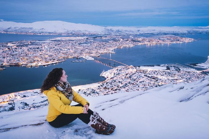 girl sitting on the snow on the top of mountain with the view of tromso city and tromso bridge in the back the girl is dressed in winter clothing