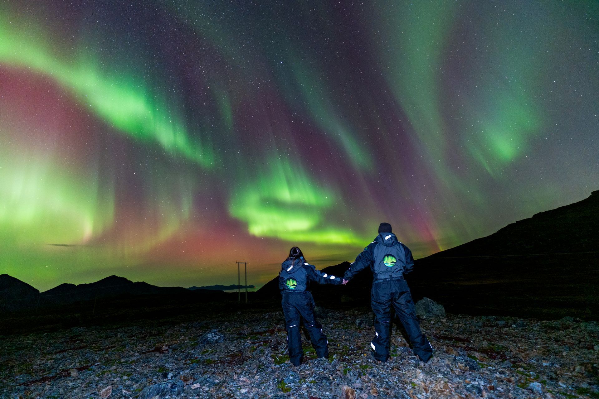 Two people standing in an Arctic landscape watching green and purple Northern Lights across the night sky in Tromsø, photographed under real conditions without edits