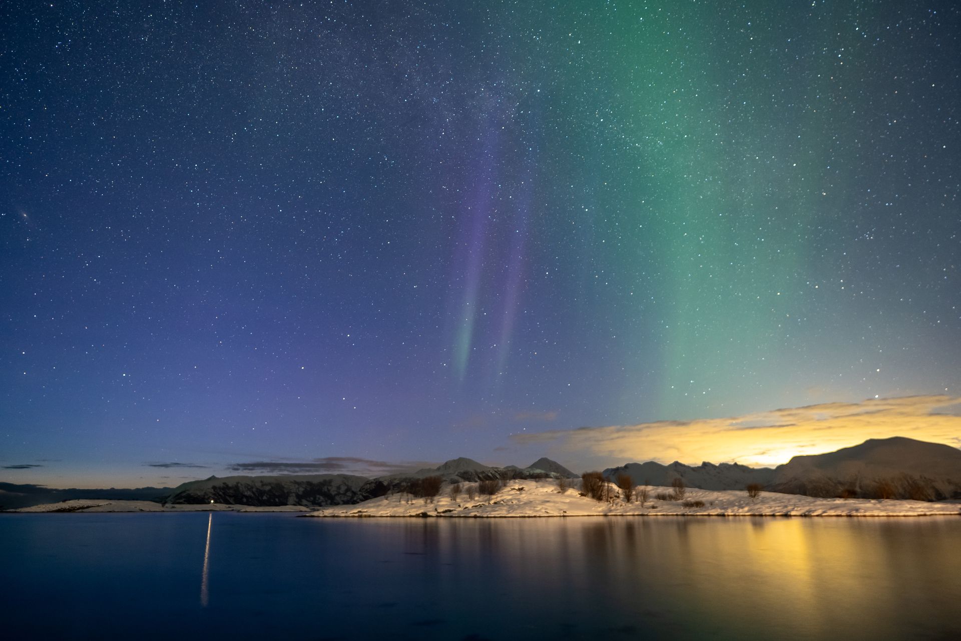 Faint green and purple Northern Lights above a calm Arctic coastline with snow-covered land and starry sky in Tromsø, photographed under real conditions without edits