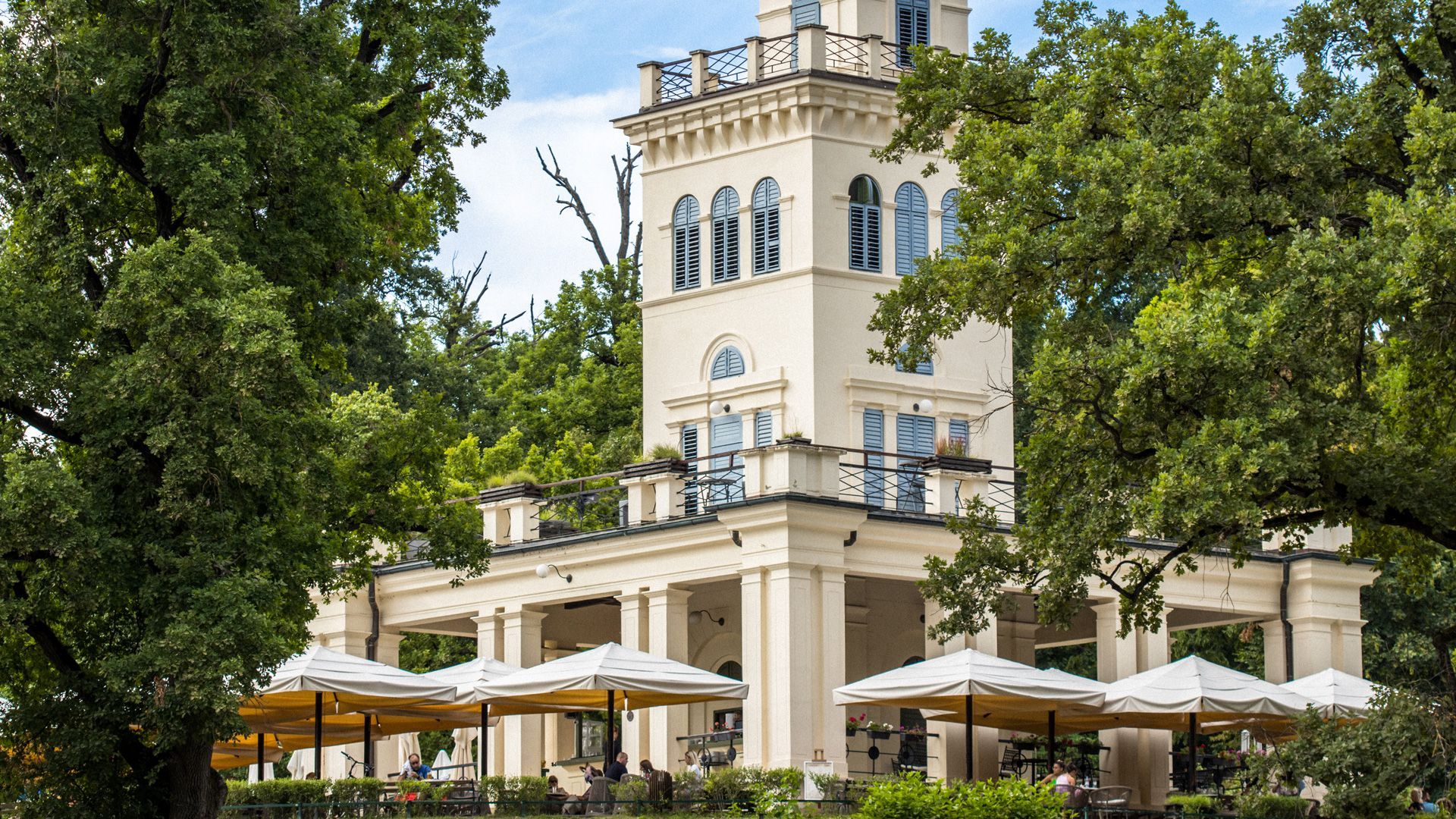 Un grande edificio bianco con una torre dell orologio è circondato da alberi e ombrelloni.