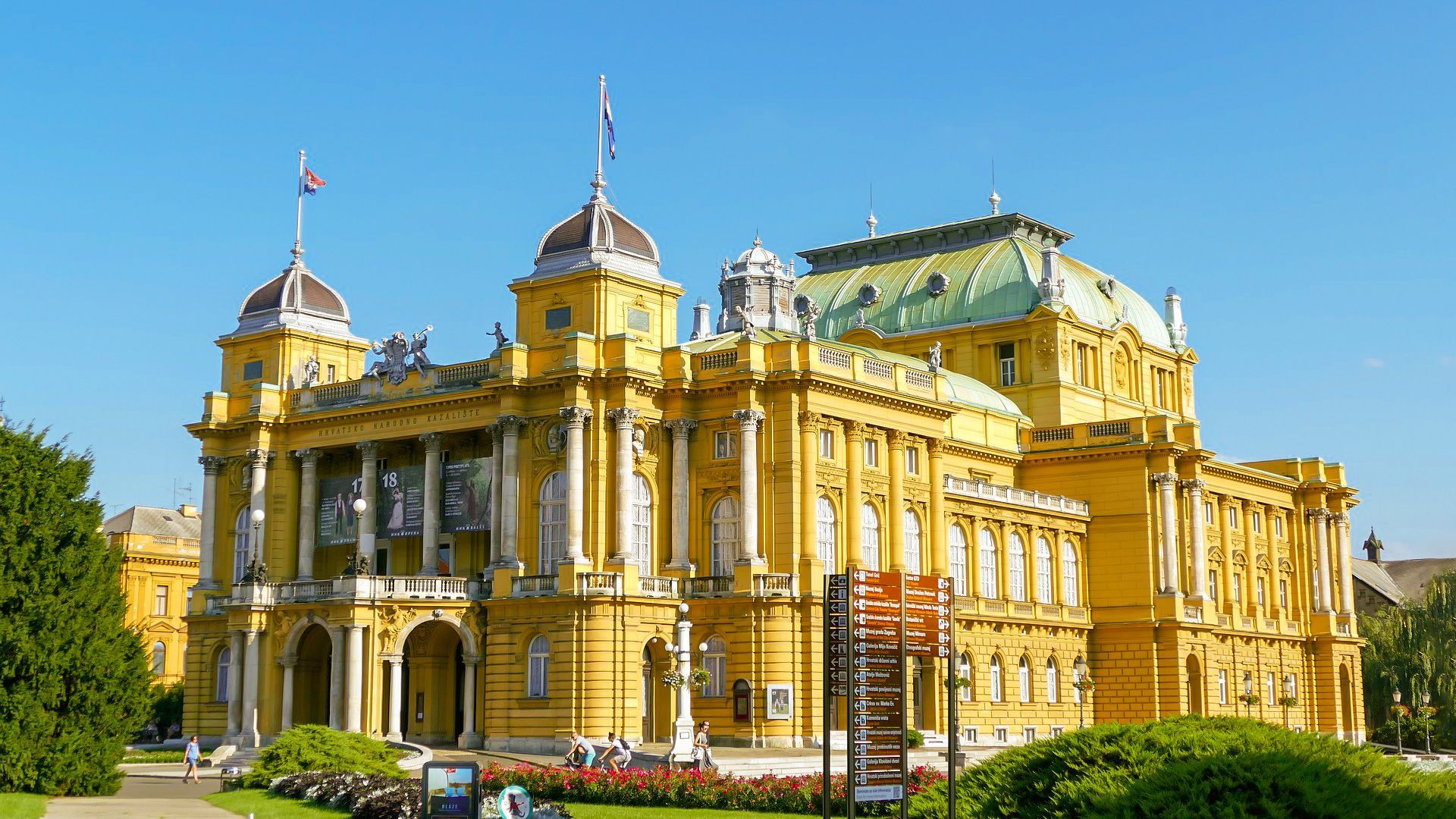 Un grande edificio giallo con il tetto verde è circondato da alberi e cespugli.