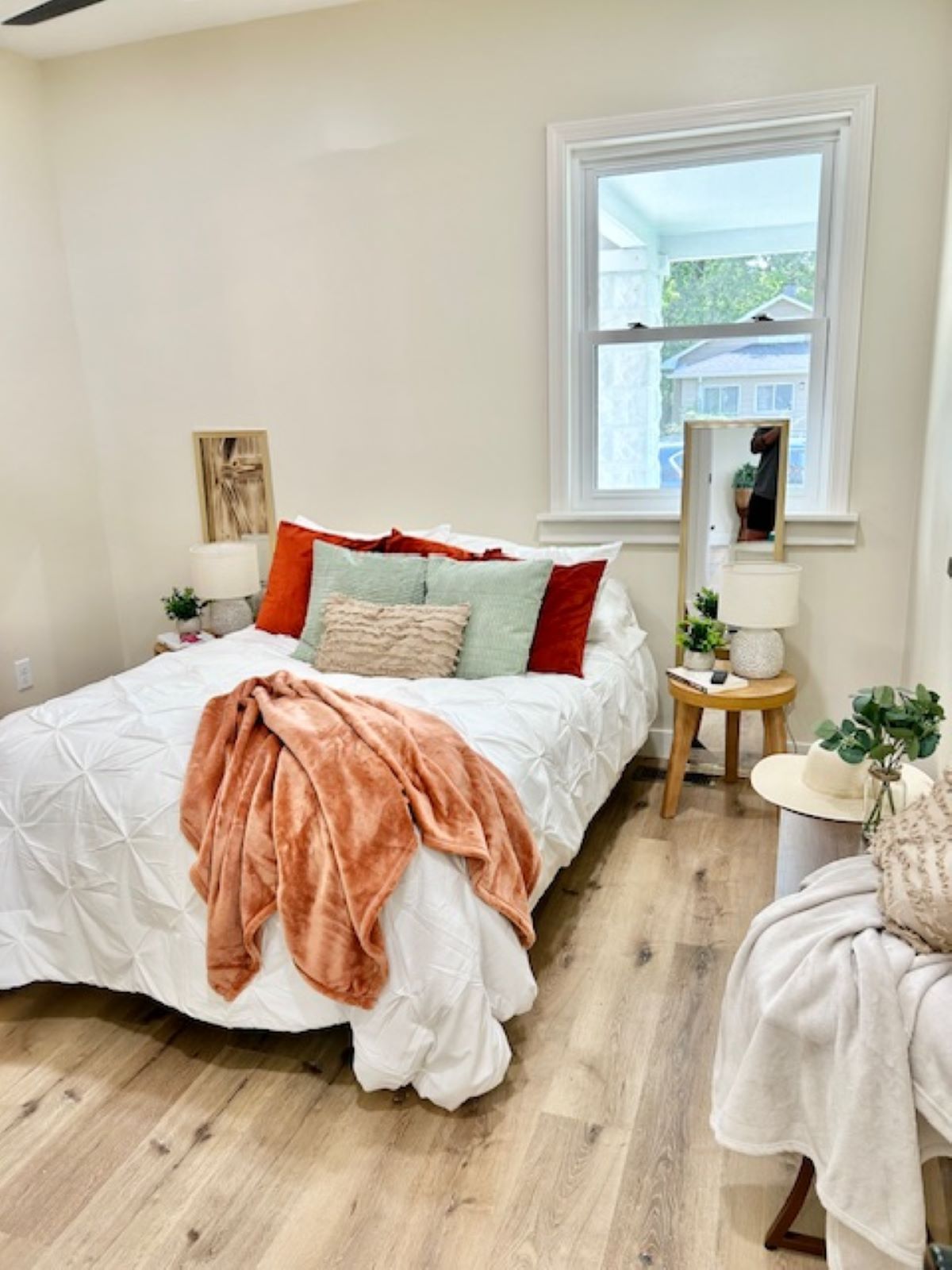 Bedroom with bed, pillows, blanket, side table with mirror, and window. Light wood flooring and neutral walls.