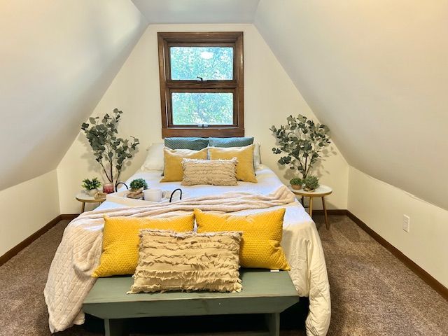 Cozy attic bedroom with a bed, yellow pillows, and a wooden bench. Natural light streams through the window.