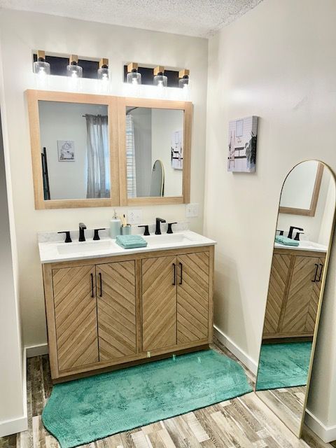 Bathroom with double vanity, light wood cabinets, two mirrors, black faucets, and a turquoise rug.