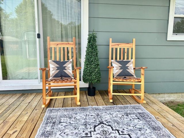 Two wooden rocking chairs with patterned pillows on a wooden deck with a rug and a tall, green plant.