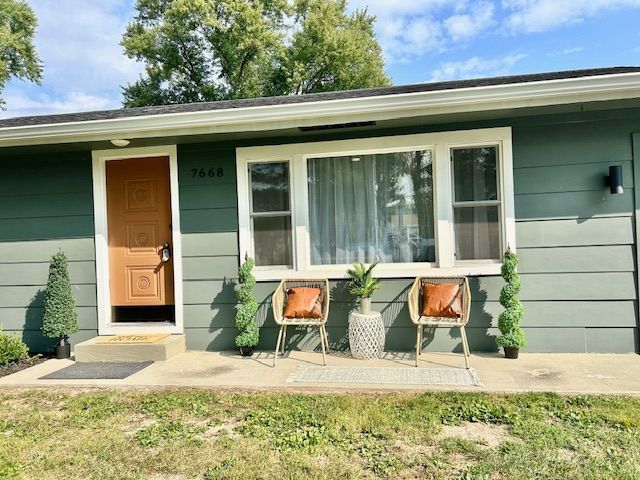 Green house exterior with brown door, window, two chairs, and topiary plants.