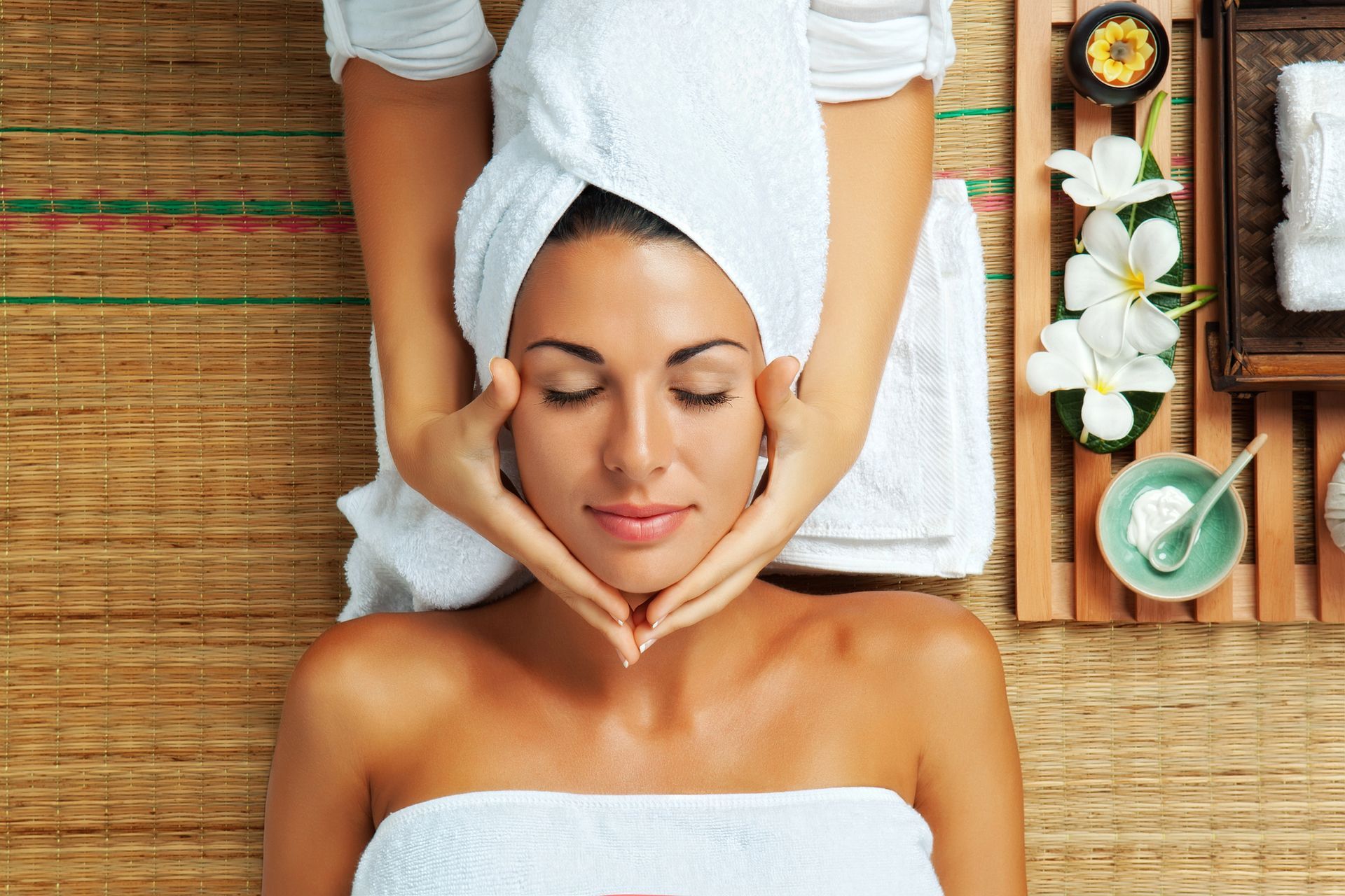 Una persona riceve un massaggio viso in una spa, avvolta in un asciugamano bianco e sdraiata su un tappetino dalla superficie ruvida, con fiori e crema nelle vicinanze.