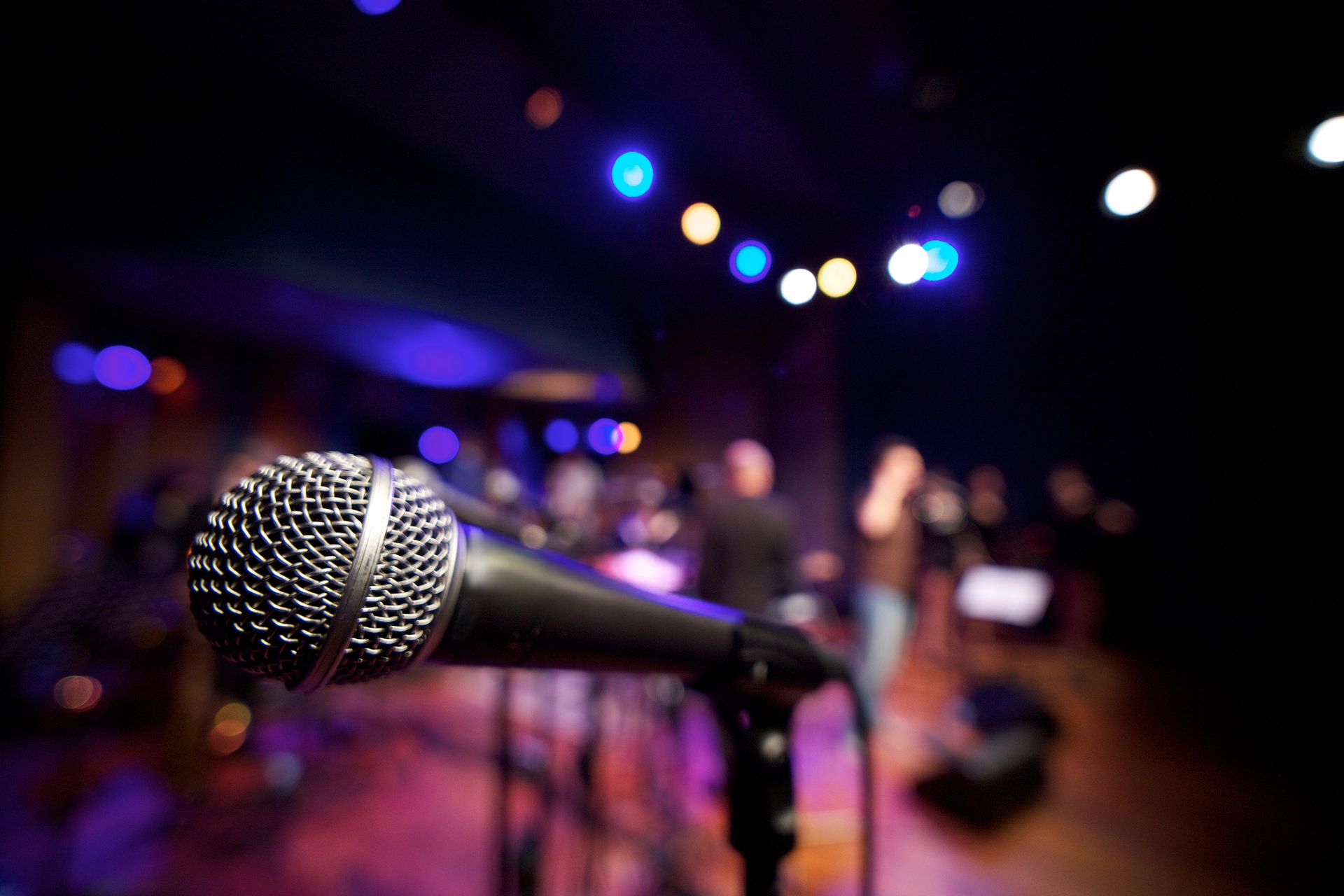 Primo piano di un microfono da palco, con una band e le luci colorate del palco sfocate sullo sfondo.