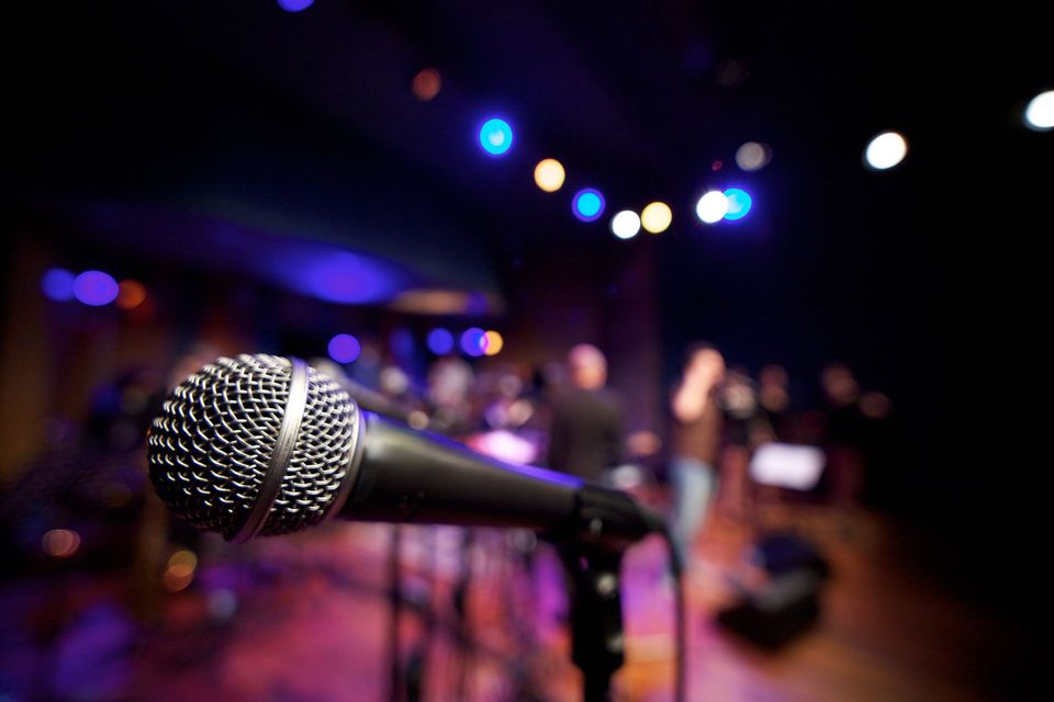 Primo piano di un microfono da palco, con una band e le luci colorate del palco sfocate sullo sfondo.
