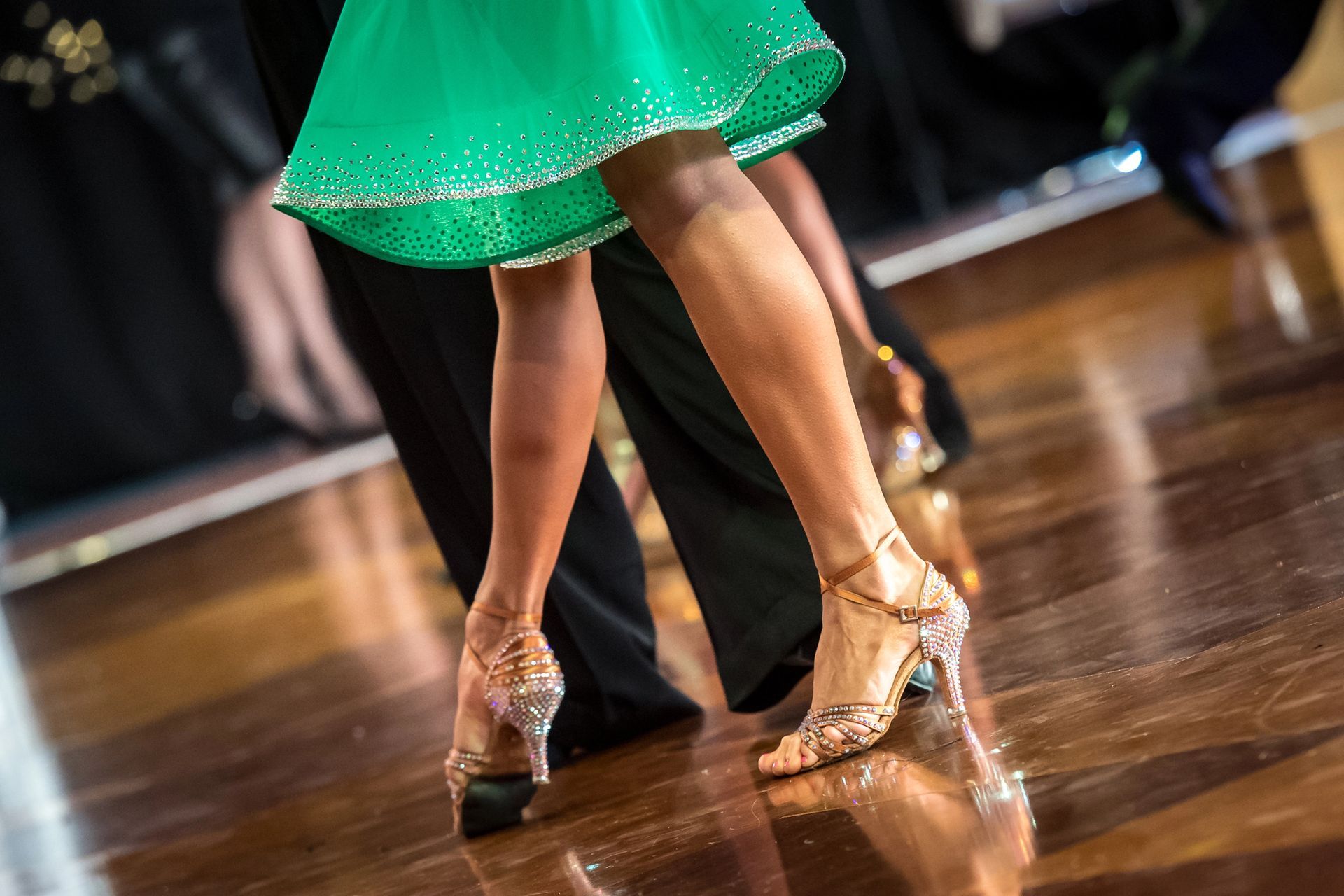 Gambe e piedi di ballerini, calzati con scarpe da ballo da sala, si muovono su un pavimento di legno lucido, indossando una gonna verde brillante.