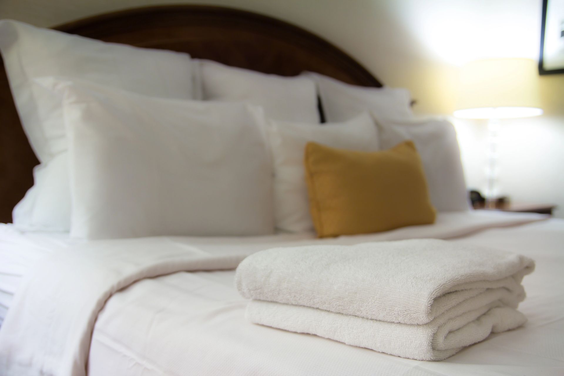 Un letto d'albergo ben rifatto con lenzuola bianche, diversi cuscini bianchi, un cuscino decorativo giallo e asciugamani bianchi piegati.