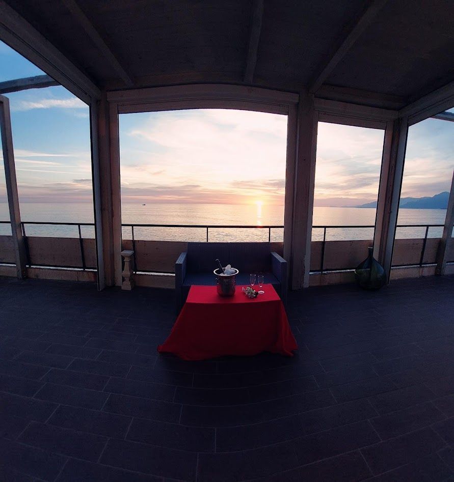 Un tavolino con una tovaglia rossa e un secchiello per il vino è posizionato davanti a un divano su un balcone che si affaccia sull'oceano al tramonto.