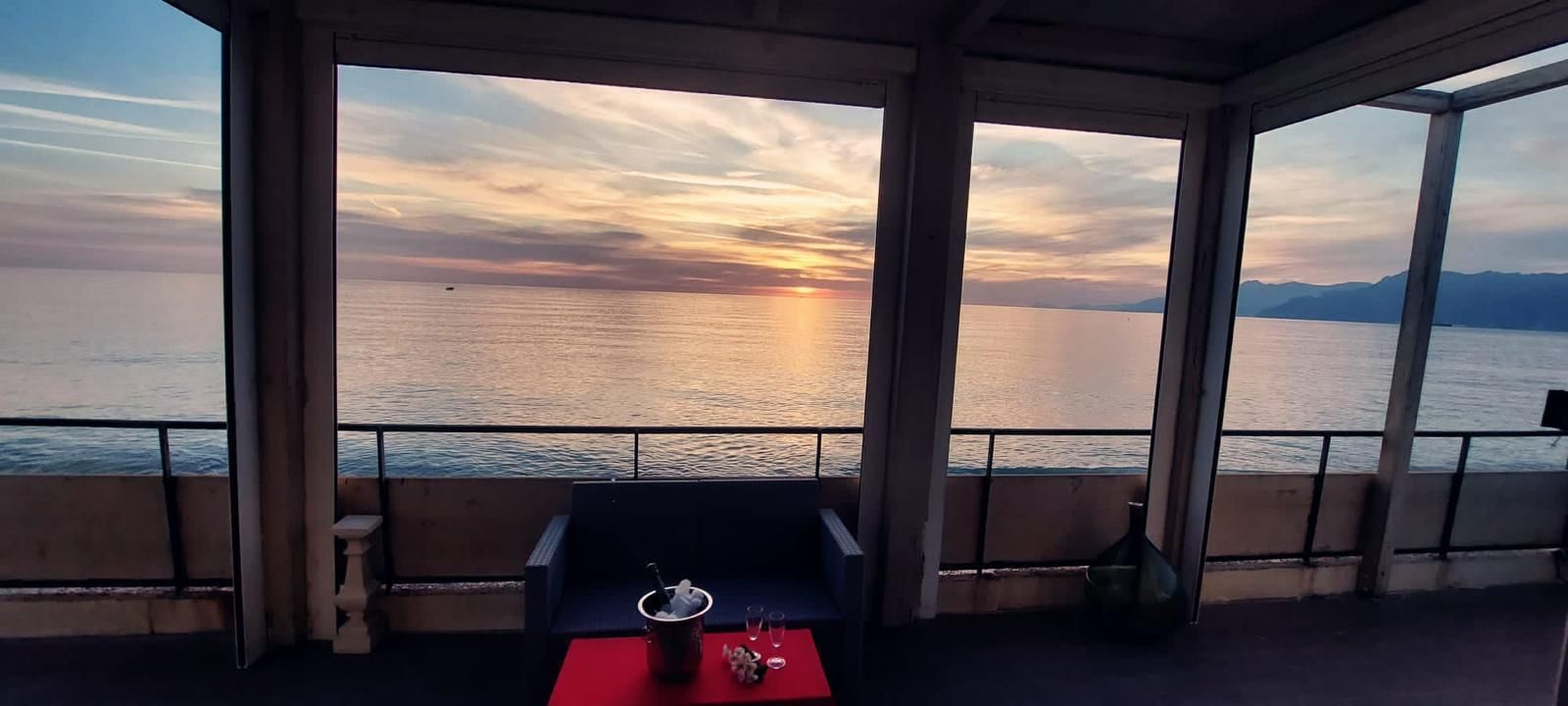 Un tramonto sul mare visto da un balcone ombreggiato con mobili scuri e un tavolo rosso.