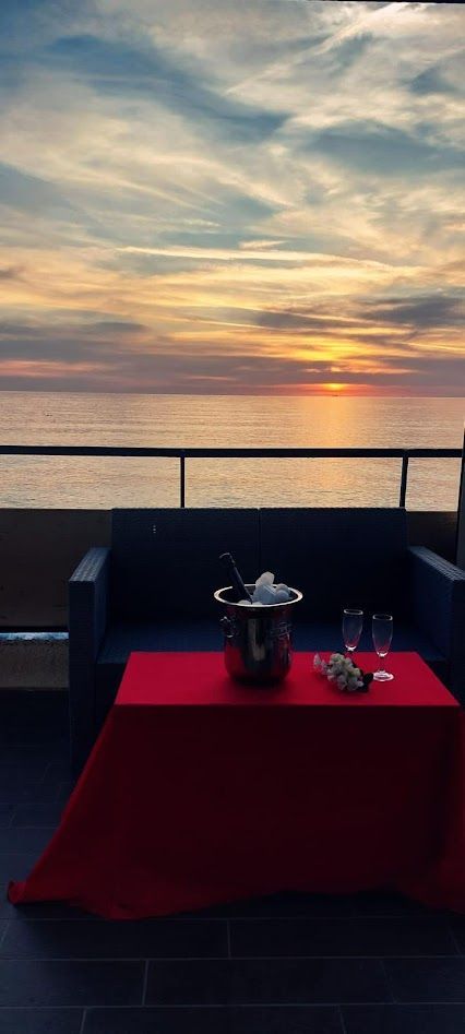 Un tramonto sull'oceano visto da un balcone con un tavolino apparecchiato con una tovaglia rossa, un secchiello per il vino e due bicchieri.