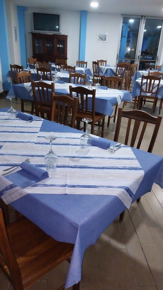 Sala da pranzo con diversi tavoli ricoperti da tovaglie a righe blu e bianche e sedie in legno, in un ambiente interno luminoso.