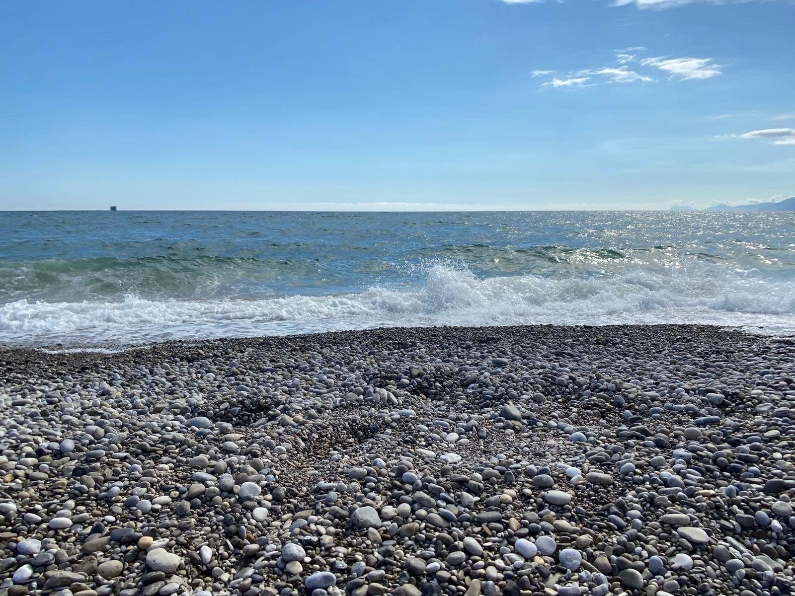 In primo piano, una spiaggia è ricoperta di piccoli ciottoli grigi che lambiscono dolci onde sotto un cielo azzurro e soleggiato.