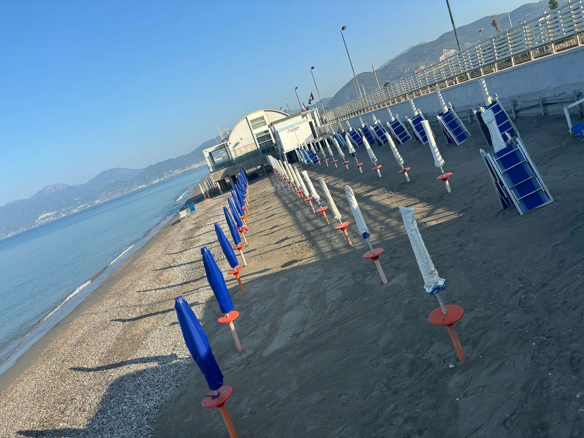 Spiaggia deserta con file di ombrelloni chiusi, blu e bianchi, allineati lungo la sabbia vicino al mare calmo e blu.