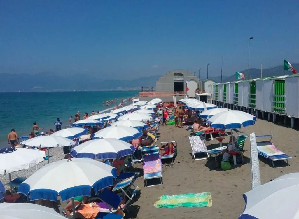 Una spiaggia affollata con file di ombrelloni a righe bianche e blu, sdraio e piccole cabine bianche in riva al mare.