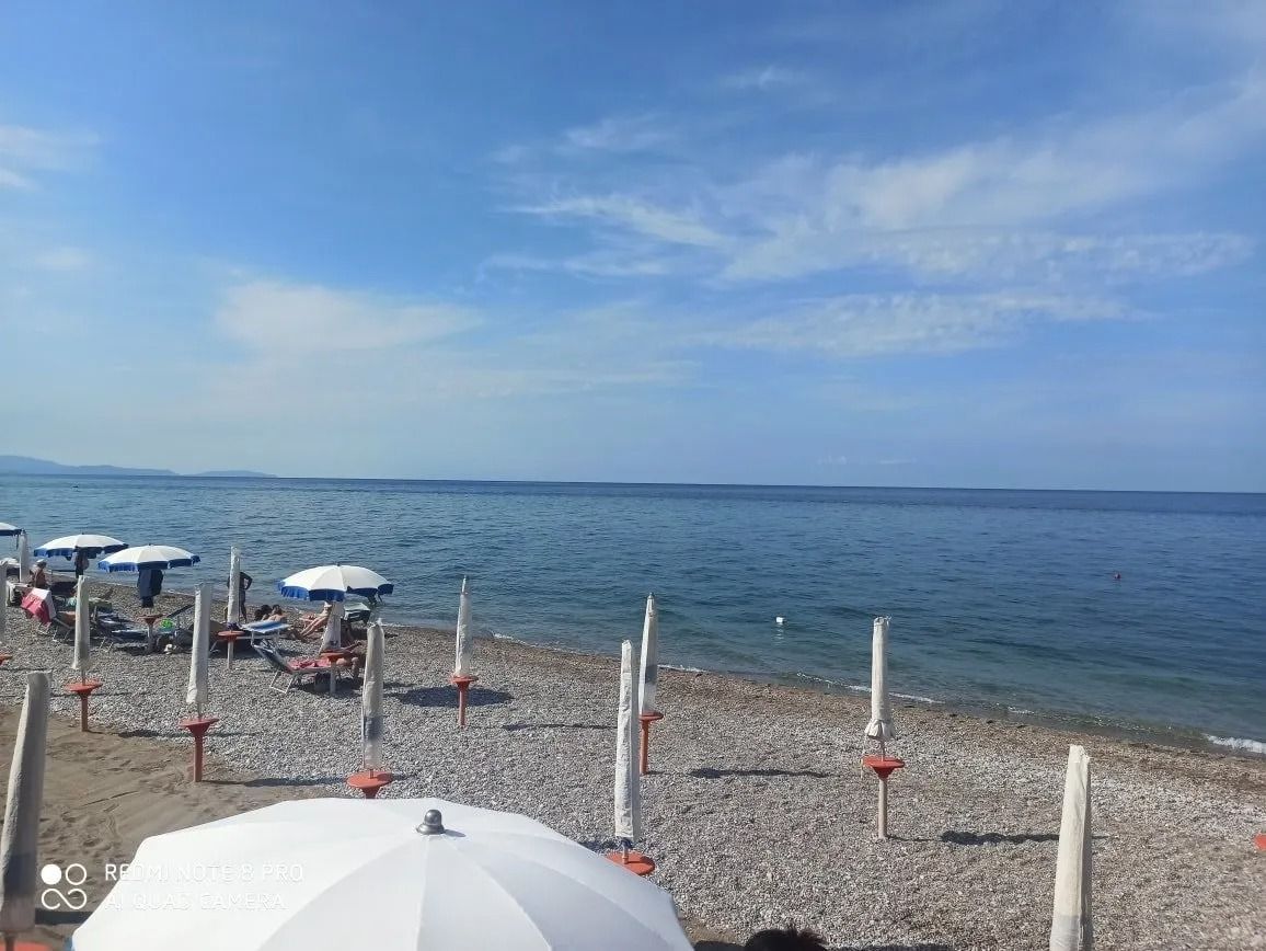 Una spiaggia di ciottoli con ombrelloni bianchi sotto un cielo azzurro, affacciata su un mare calmo.