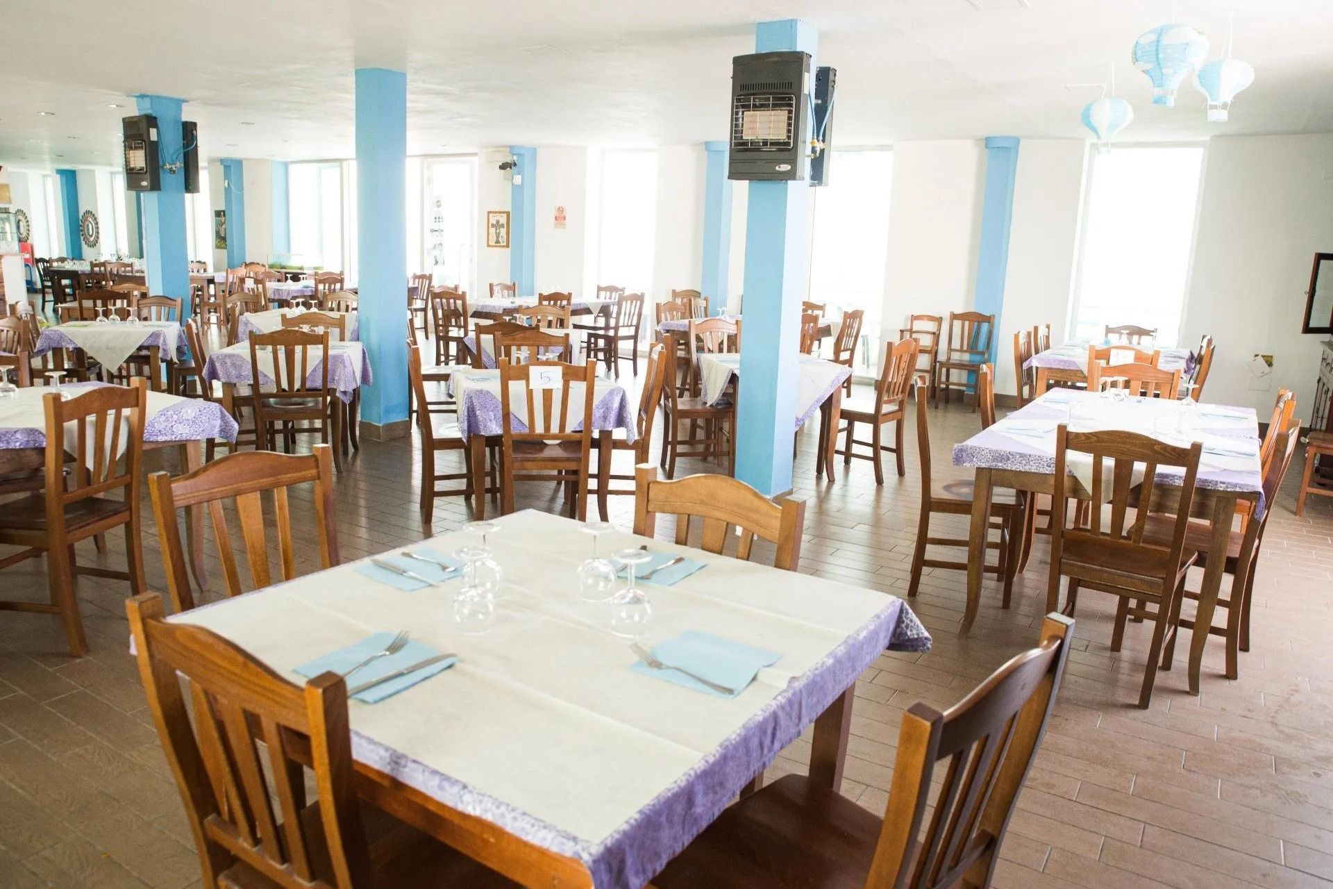Una sala da pranzo luminosa e spaziosa, con diversi tavoli apparecchiati con tovaglie bianche, sedie in legno e dettagli blu sui pilastri.