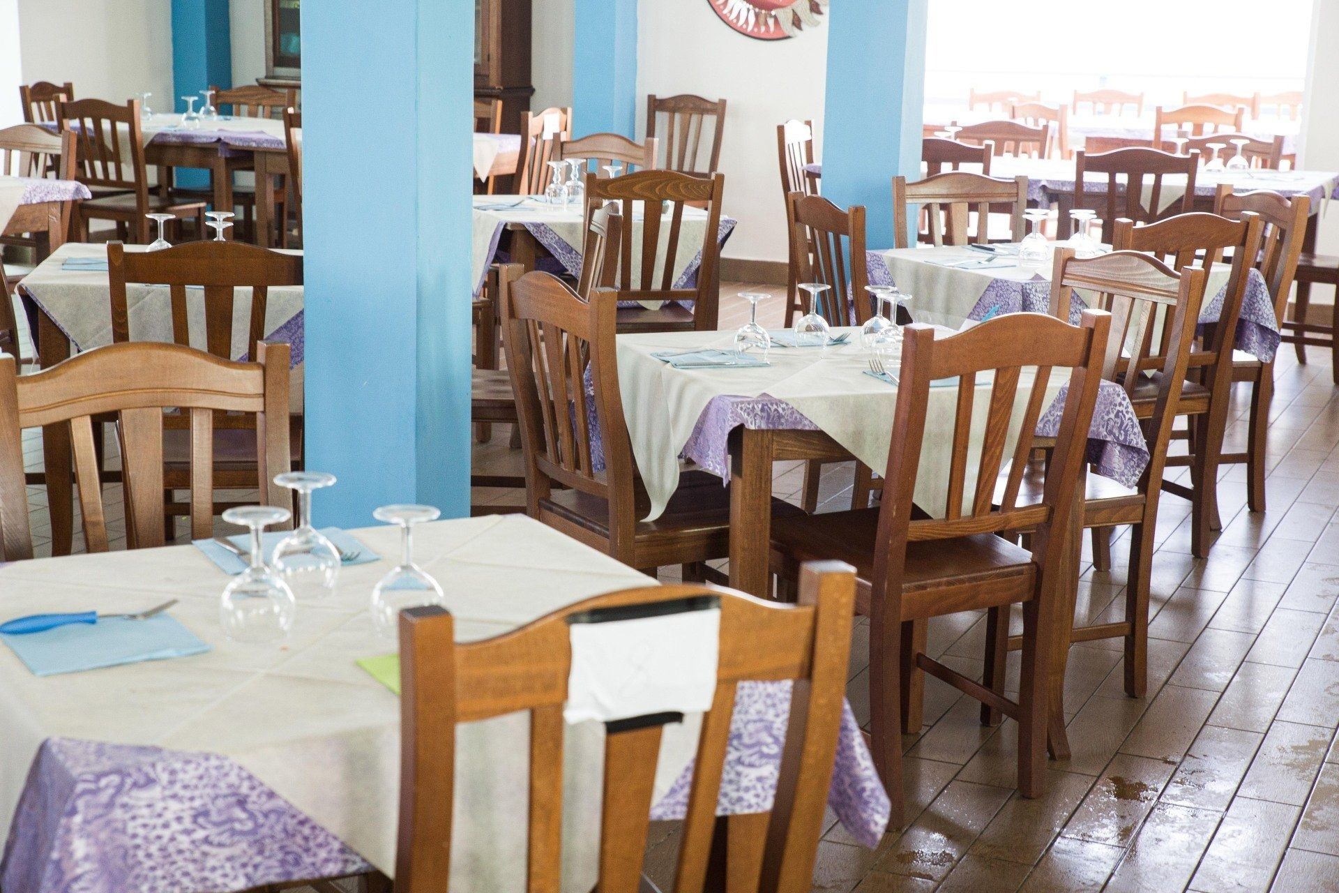 Tavoli vuoti in un ristorante con sedie di legno, tovaglie chiare e colonne blu in una sala da pranzo luminosa con pavimento in legno.