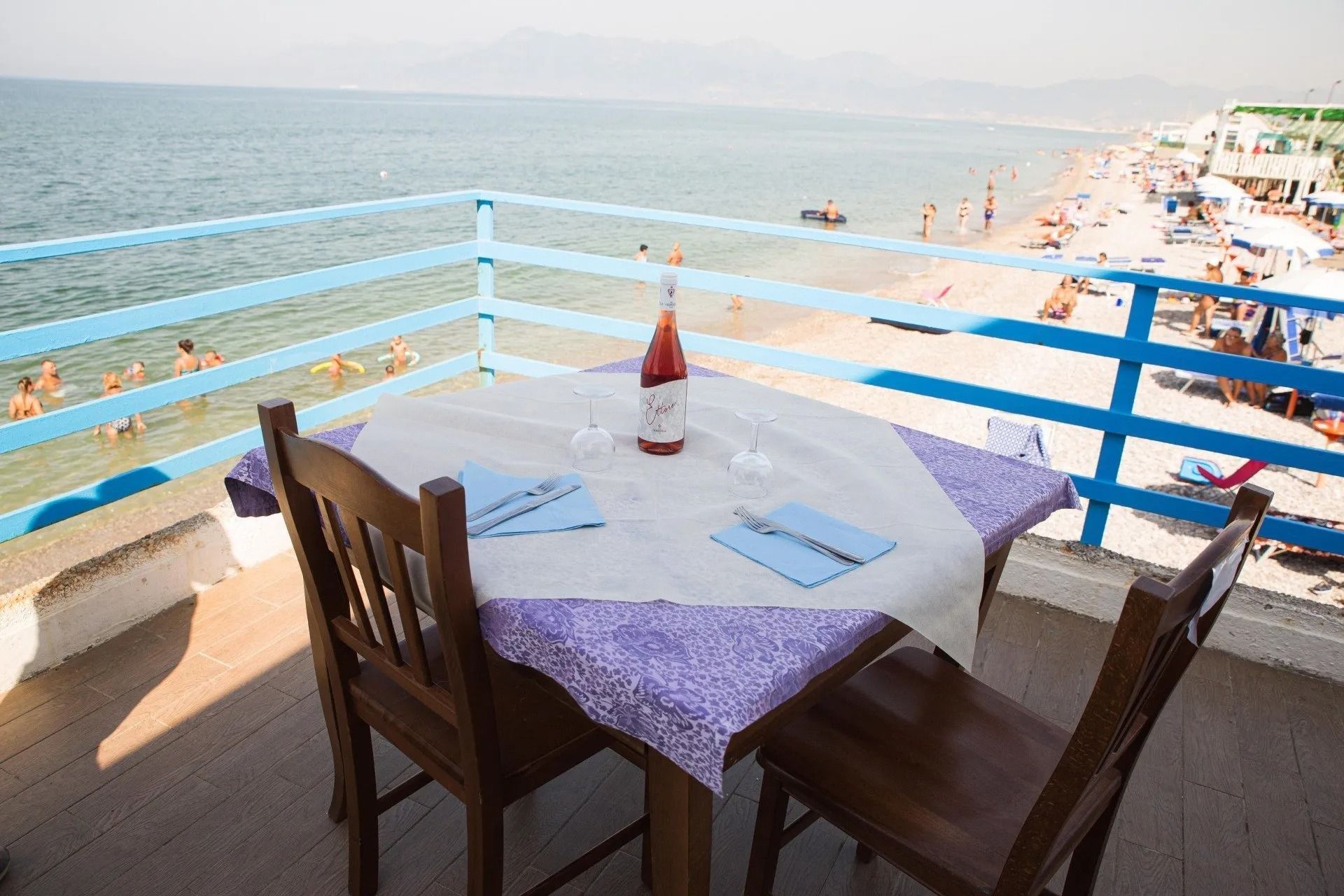 Un tavolo apparecchiato per due su un balcone con vista sulla spiaggia, con una bottiglia di vino su una tovaglia e ringhiere blu.