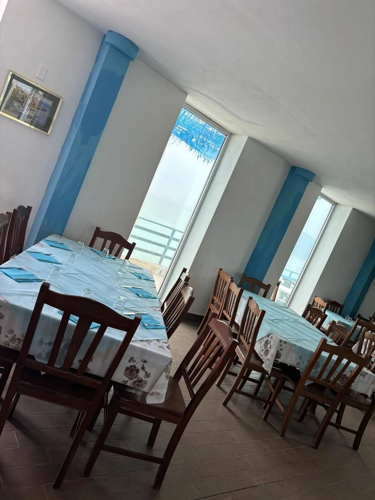 La sala da pranzo di un ristorante con colonne blu, tavoli chiari e sedie in legno scuro, affacciata su una vista sull'oceano.