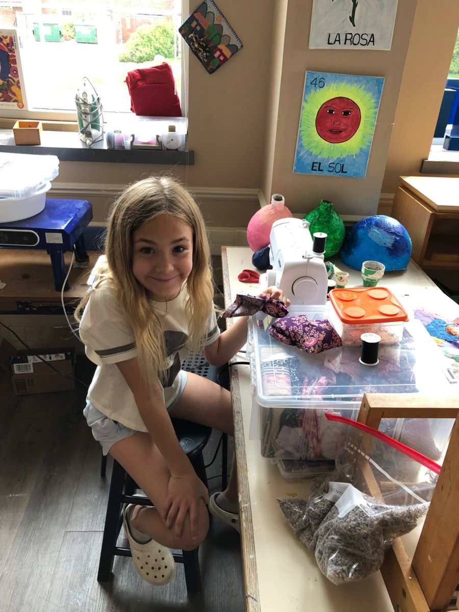 A girls sitting at a table with a sawing machine and cloths in her hand, smiling at the camara 