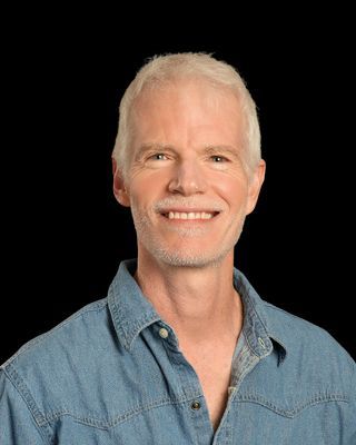 A man in a blue shirt is smiling for the camera in a park.