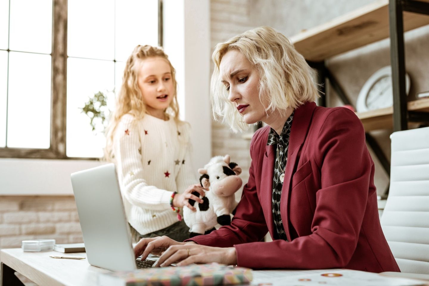 Child showing stuffed cow to mother in red blazer works while she works on a laptop.