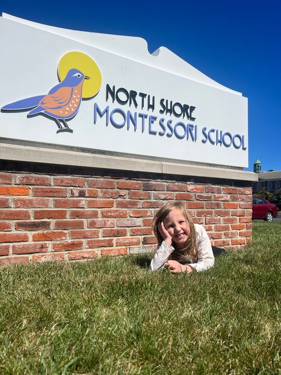 A little girl is laying in the grass in front of a north shore montessori school sign.