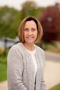 A woman in a grey sweater and white shirt is smiling for the camera.