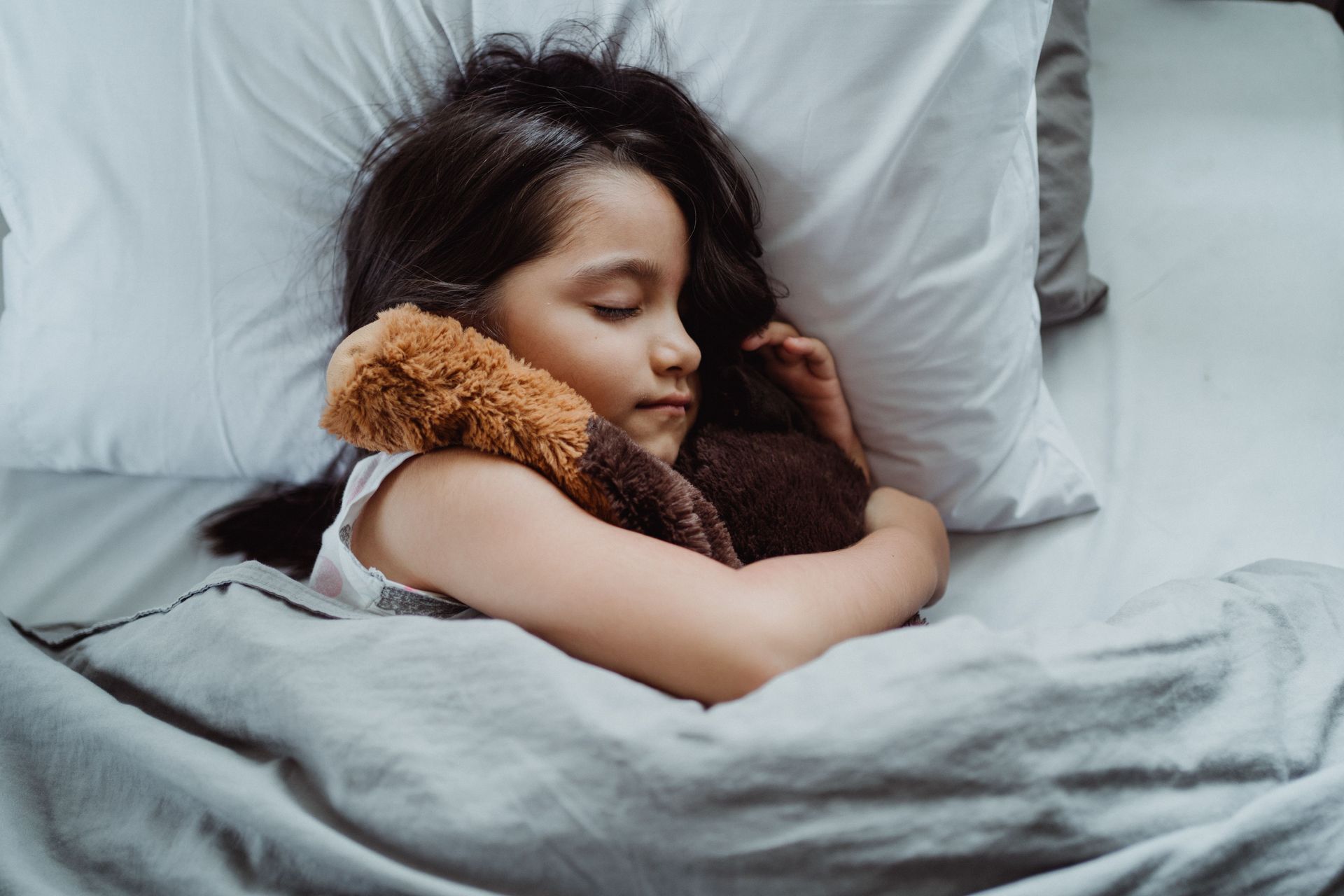 Child sleeping in bed, hugging a brown stuffed animal under gray blankets.