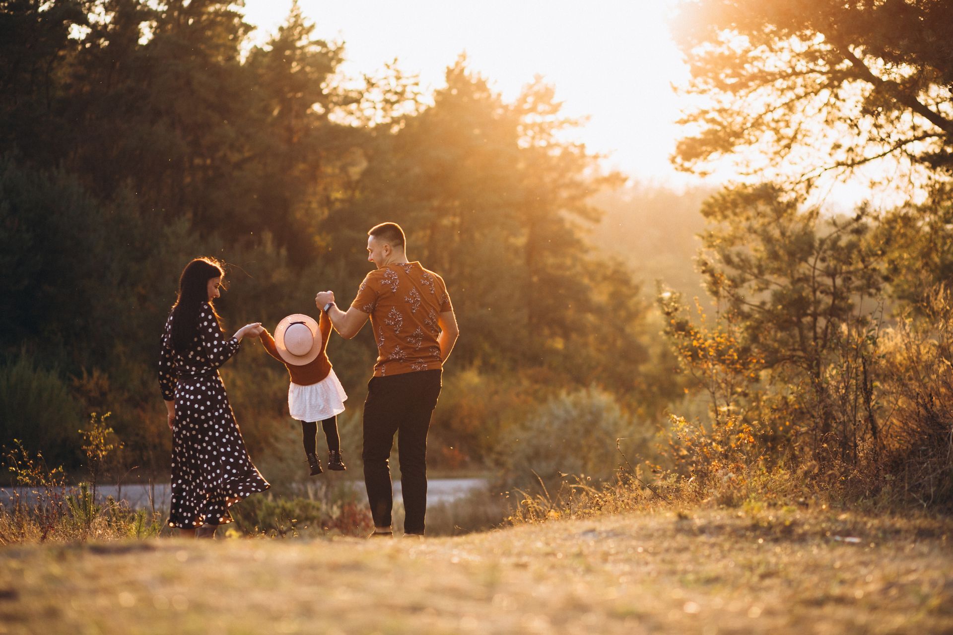 Parents swinging a child in a sunlit forest. The child wears a hat and is airborne; parents are looking at the child.