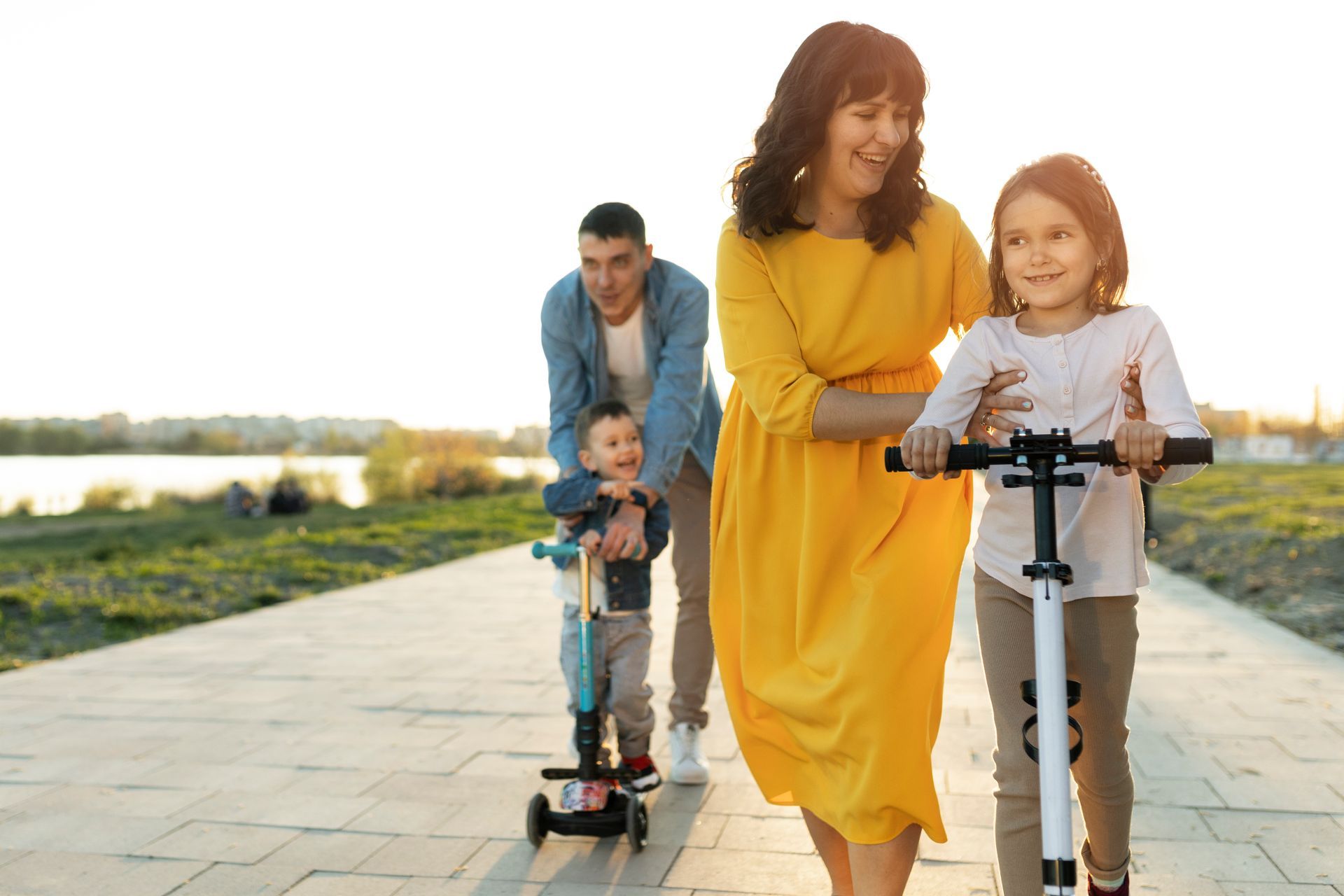 Family rides scooters on a paved path near water. Smiling mother guides daughter, father assists toddler.