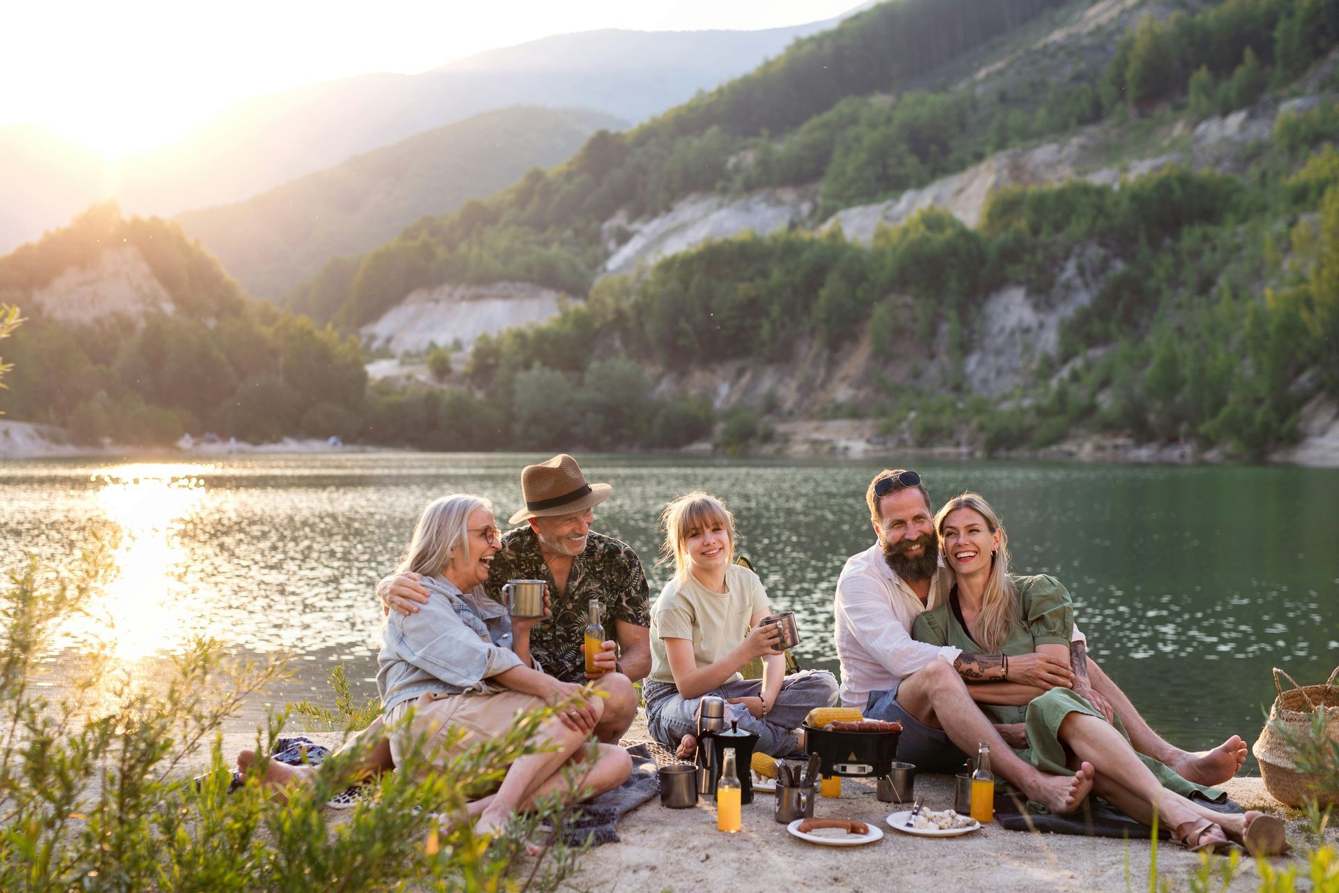 Family picnic by a lake: people sitting on a blanket, mountains and trees in background.