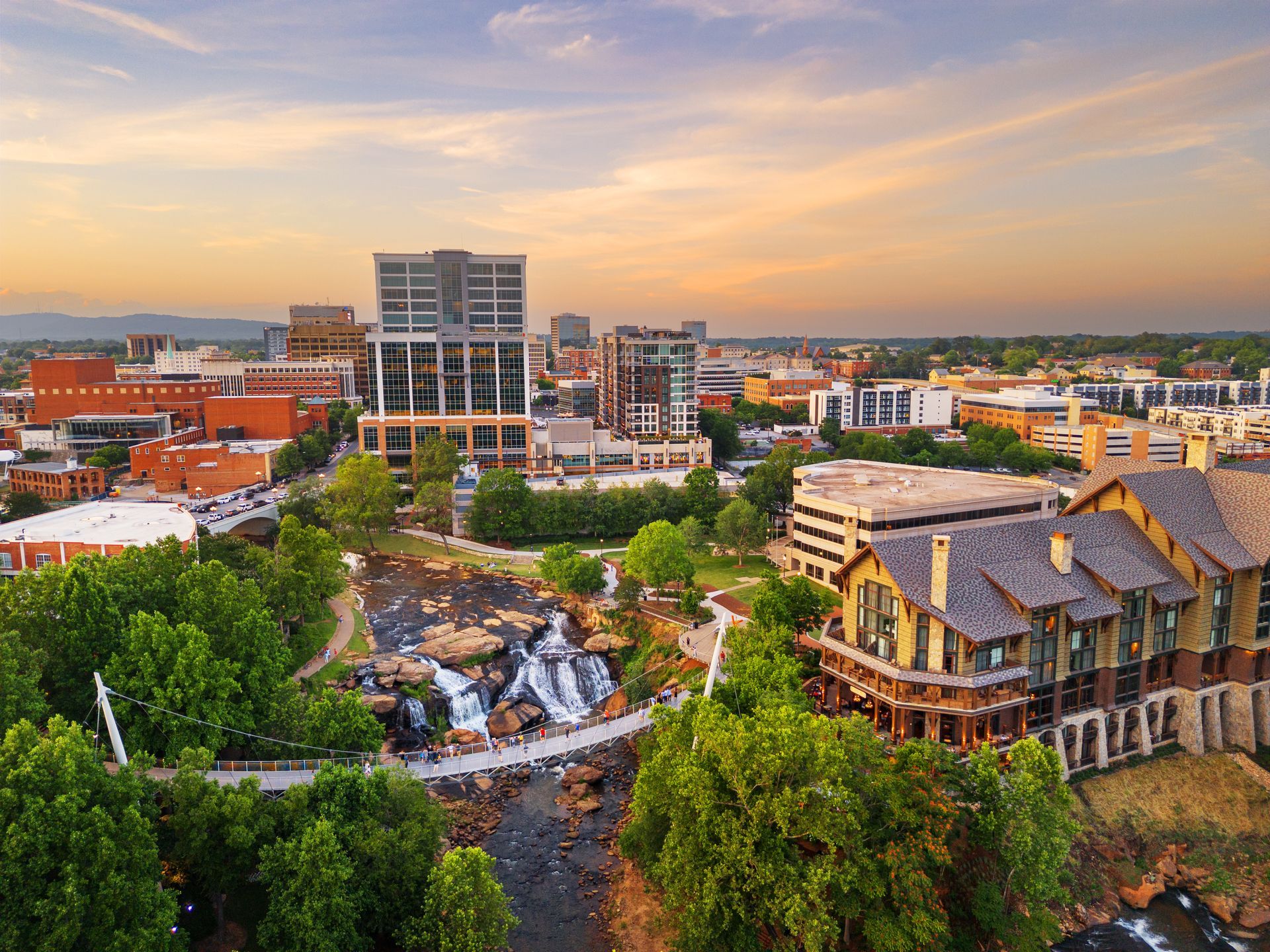 City skyline with buildings, waterfall, and bridge under a sunset sky.