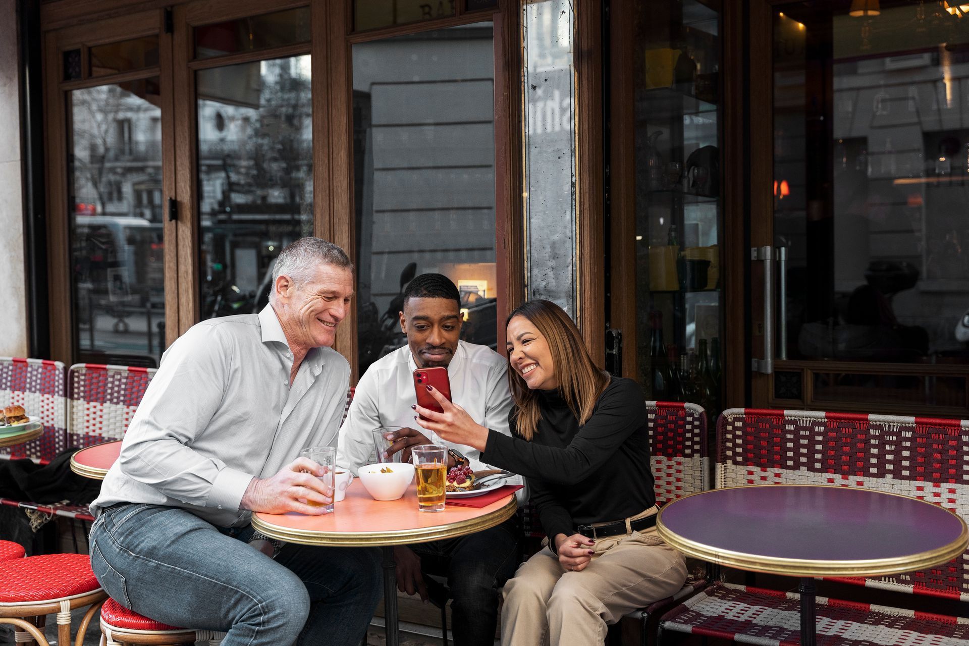 Three people at an outdoor cafe looking at a phone. The woman points at the screen, smiling.