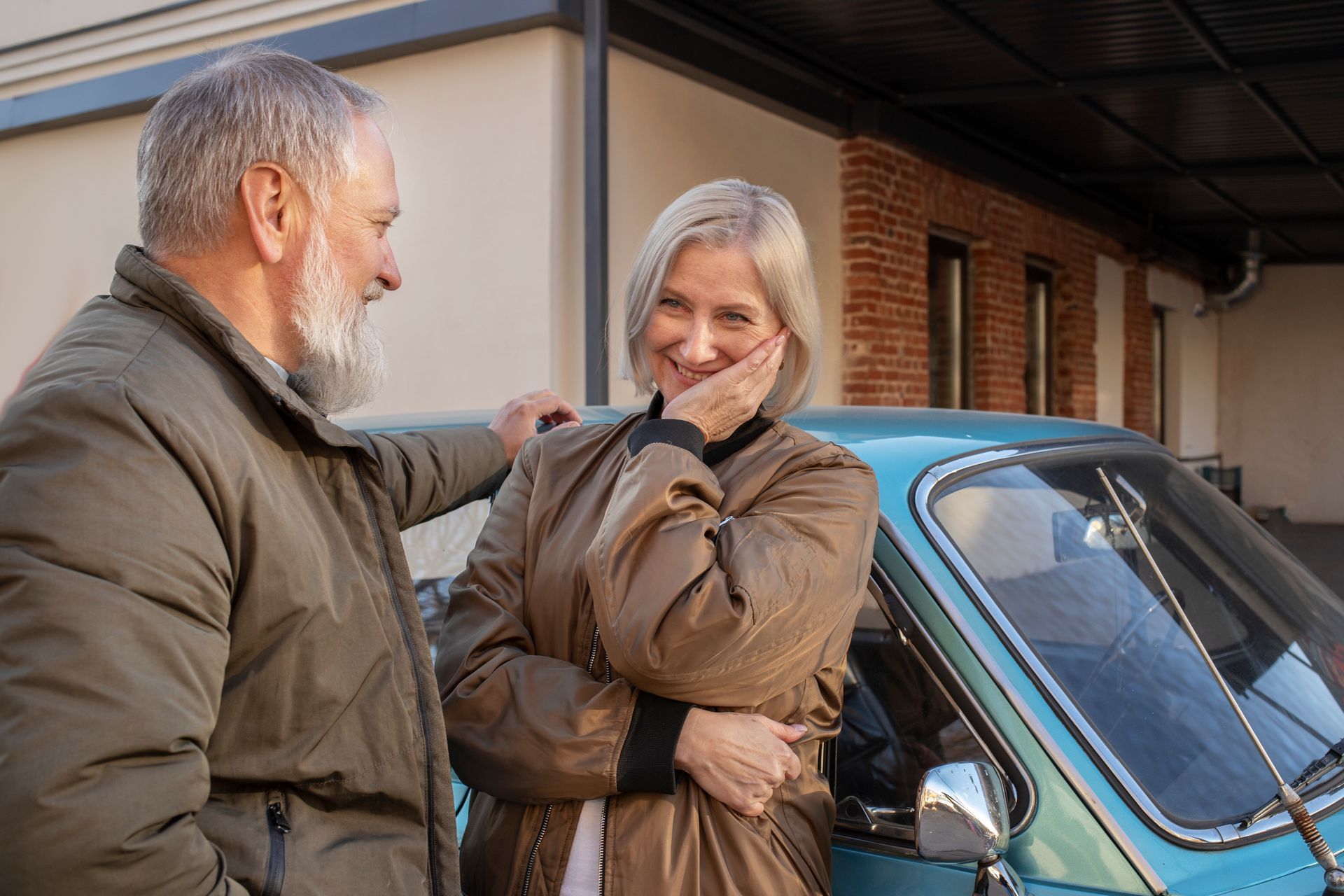 Elderly couple smiling near a turquoise car outdoors; the woman touches her face while the man leans on the car.