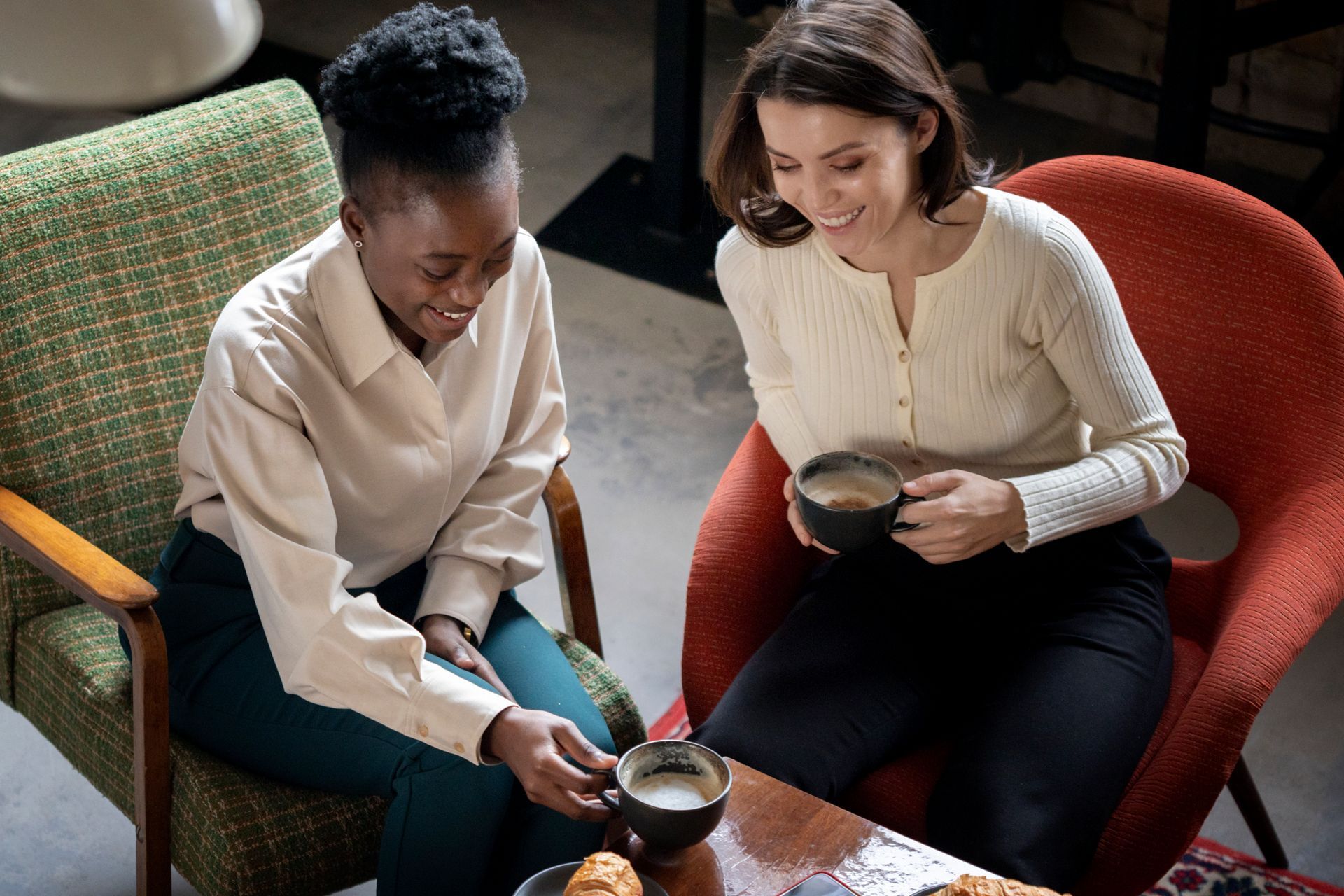 Two people, one in a light beige shirt and one in a cream-colored cardigan, drink coffee while laughing in armchairs.